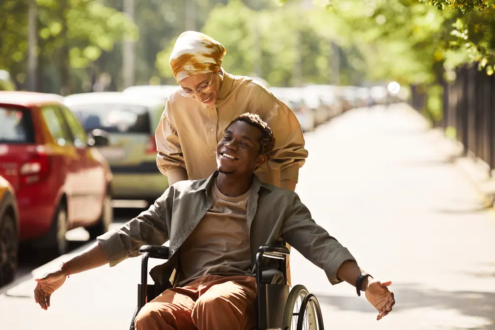 Young man I wheelchair accompanied by his female support worker. They're outside soaking the sunshine.
