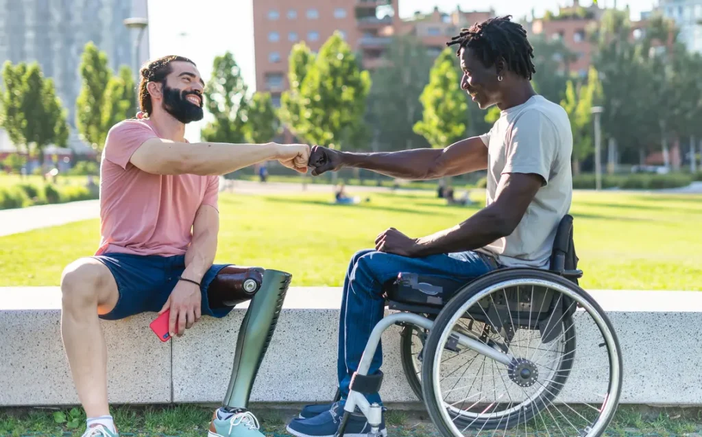 Two men, one in a wheelchair and the other with a prosthetic leg both fist bumping while sitting on a ledge at the park