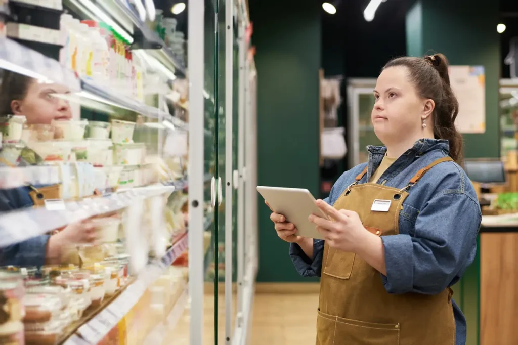 Empowered young woman with Down syndrome in her work uniform looking at a mobile tablet and items in the fridge of the grocery store where she works